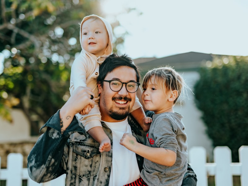 A dad laughing with his toddler on his shoulders and daughter in his arm