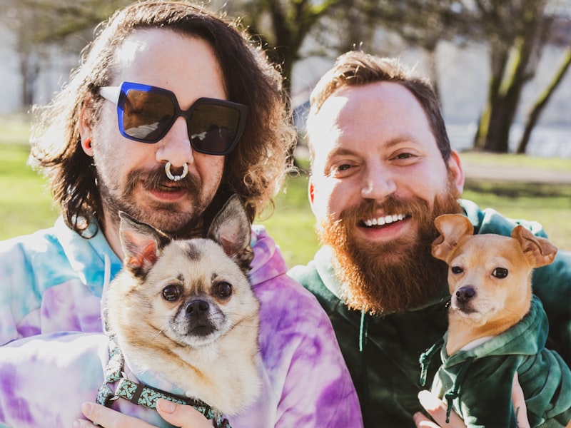 Two men smiling in the park, each holding one of their small dogs